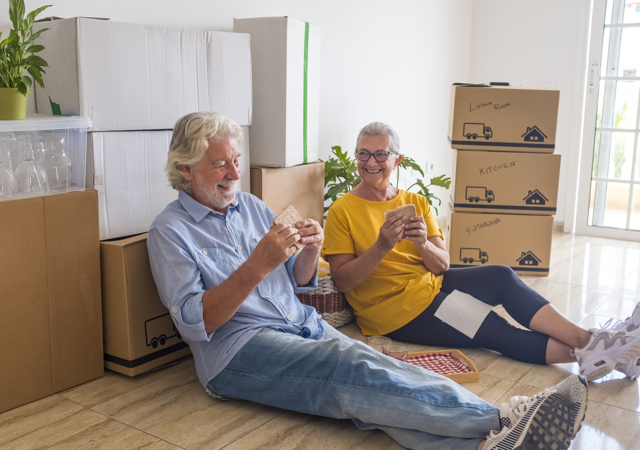 People Sitting On Floor At Home Two Young Male Movers In Uniform Placing Television In Living Room