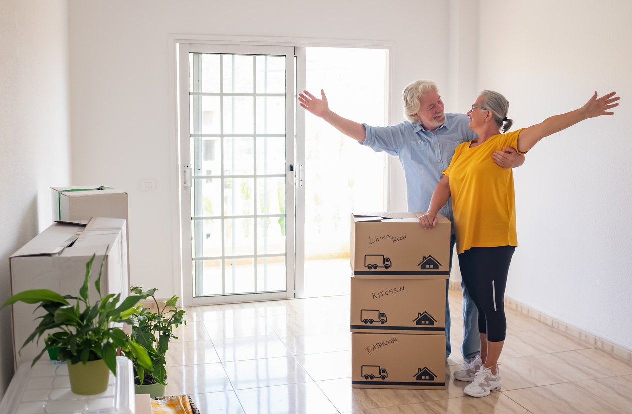 Smiling caucasian senior couple white haired open arms looking at each other's eyes during relocation, excited for a new beginning. Moving boxes on the floor, empty apartment Smiling caucasian senior couple white haired open arms looking at each other's eyes during relocation, excited for a new beginning. Moving boxes on the floor, empty apartment