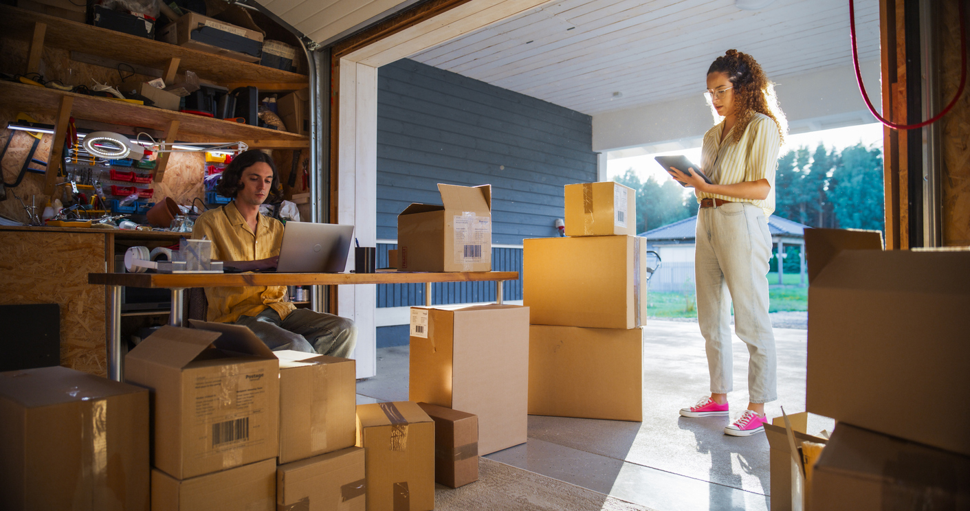 Caucasian Male Small Business Owner Using Laptop Computer In Home Garage To Fill Online Orders And Talking To Hispanic Female Manager. Multiethnic Businesswoman Using Tablet To Check Inventory. Caucasian Male Small Business Owner Using Laptop Computer In Home Garage To Fill Online Orders And Talking To Hispanic Female Manager. Multiethnic Businesswoman Using Tablet To Check Inventory.