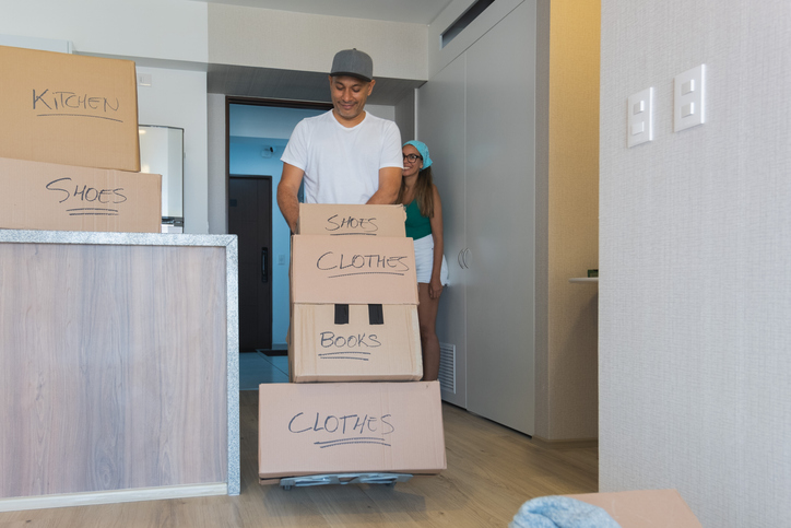 A man and a woman are moving boxes into a new apartment. New home concept. A man and a woman are moving boxes into a new apartment. The boxes are labeled with words like clothes and shoes. The man is pushing a cart with the boxes on it, while the woman is standing nearby