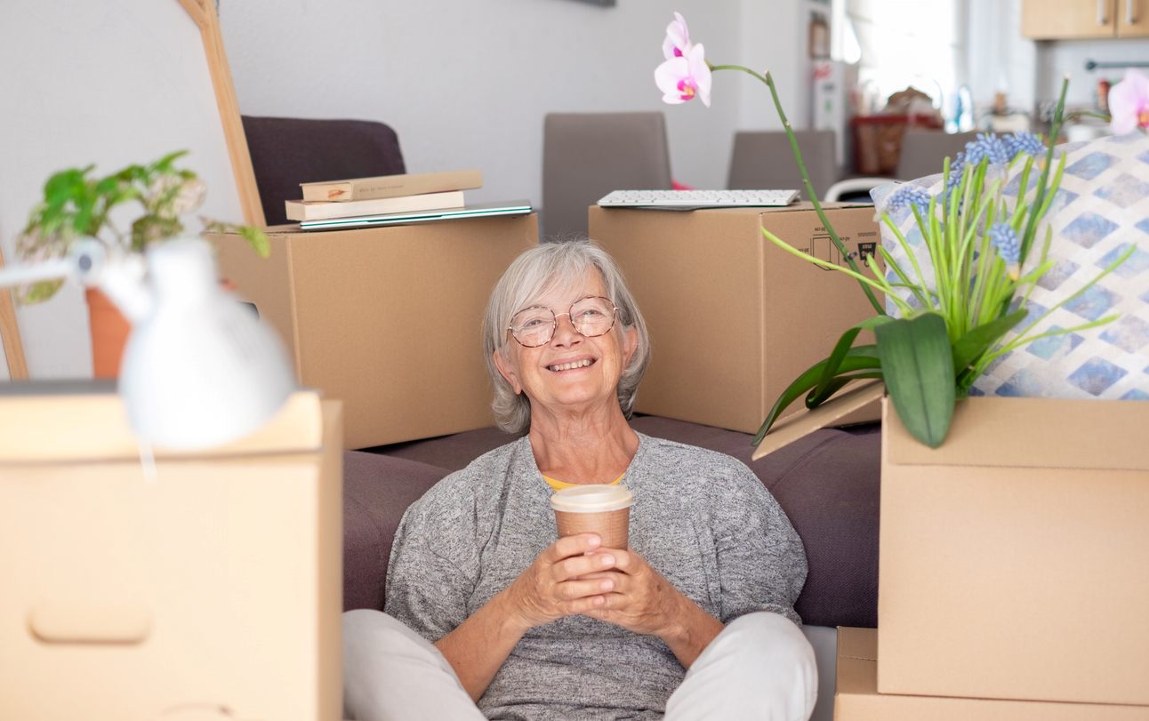 iStock-1700123847 Older woman seated on the floor with a coffee cup surrounded by moving boxes.