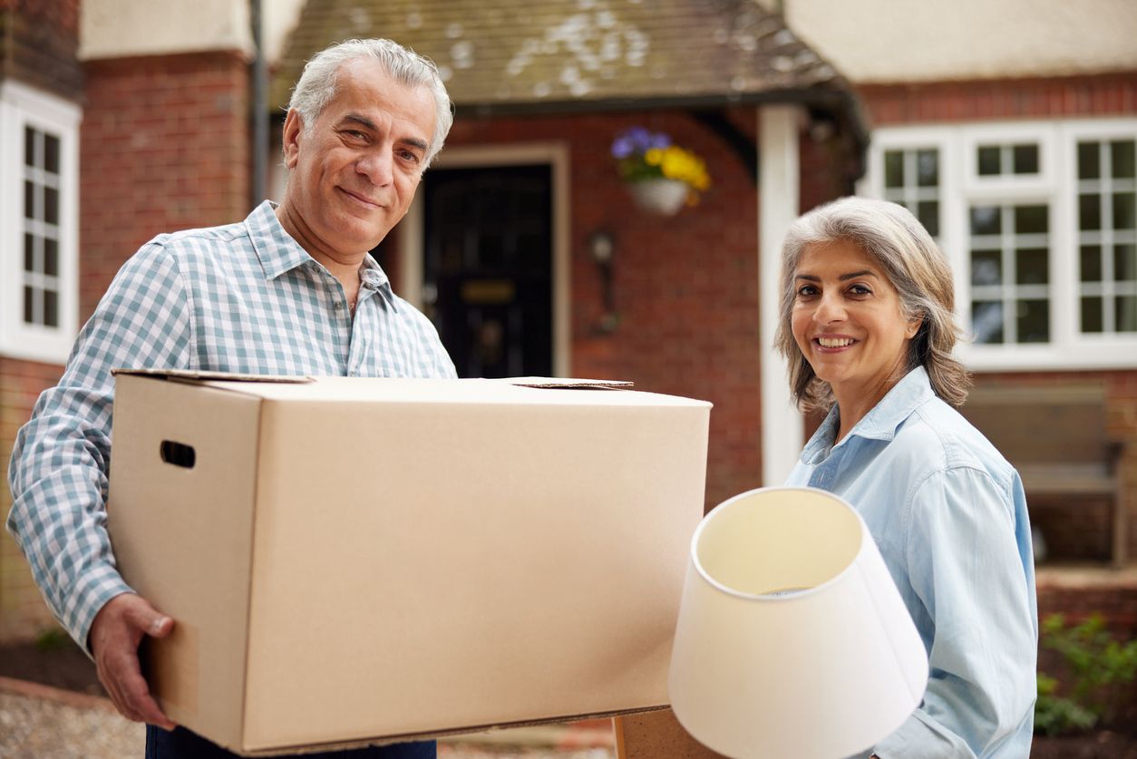 An older couple holding a box and lamp outside of a house.