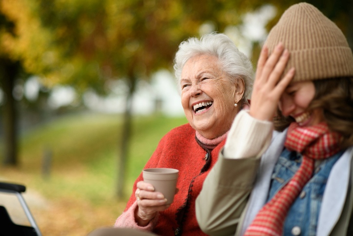 A granddaughter with her grandma sitting on a bench in autumn, laughing and drinking coffee.