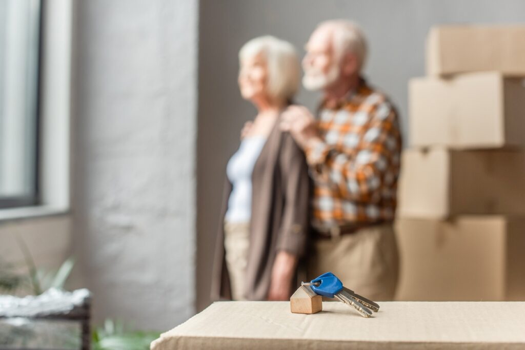 A happy elderly couple stands together in their new home, with their keys dramatically framed in the foreground.