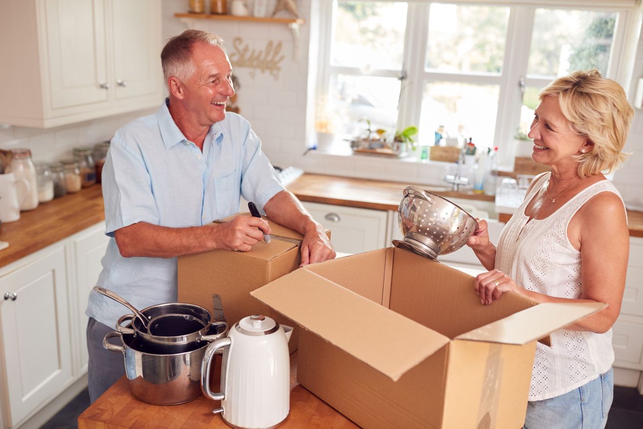 A senior couple smiles and laughs together as they pack boxes in their kitchen while downsizing.