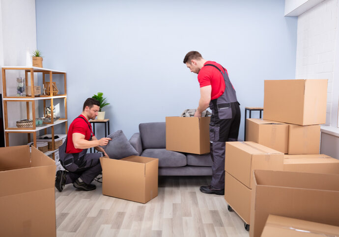 Men Loading The Cardboard Boxes During Moving Two Young Movers In Uniform Picking And Putting Products In The Cardboard Boxes