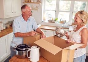 A senior couple smiles and laughs together as they pack boxes in their kitchen while downsizing.