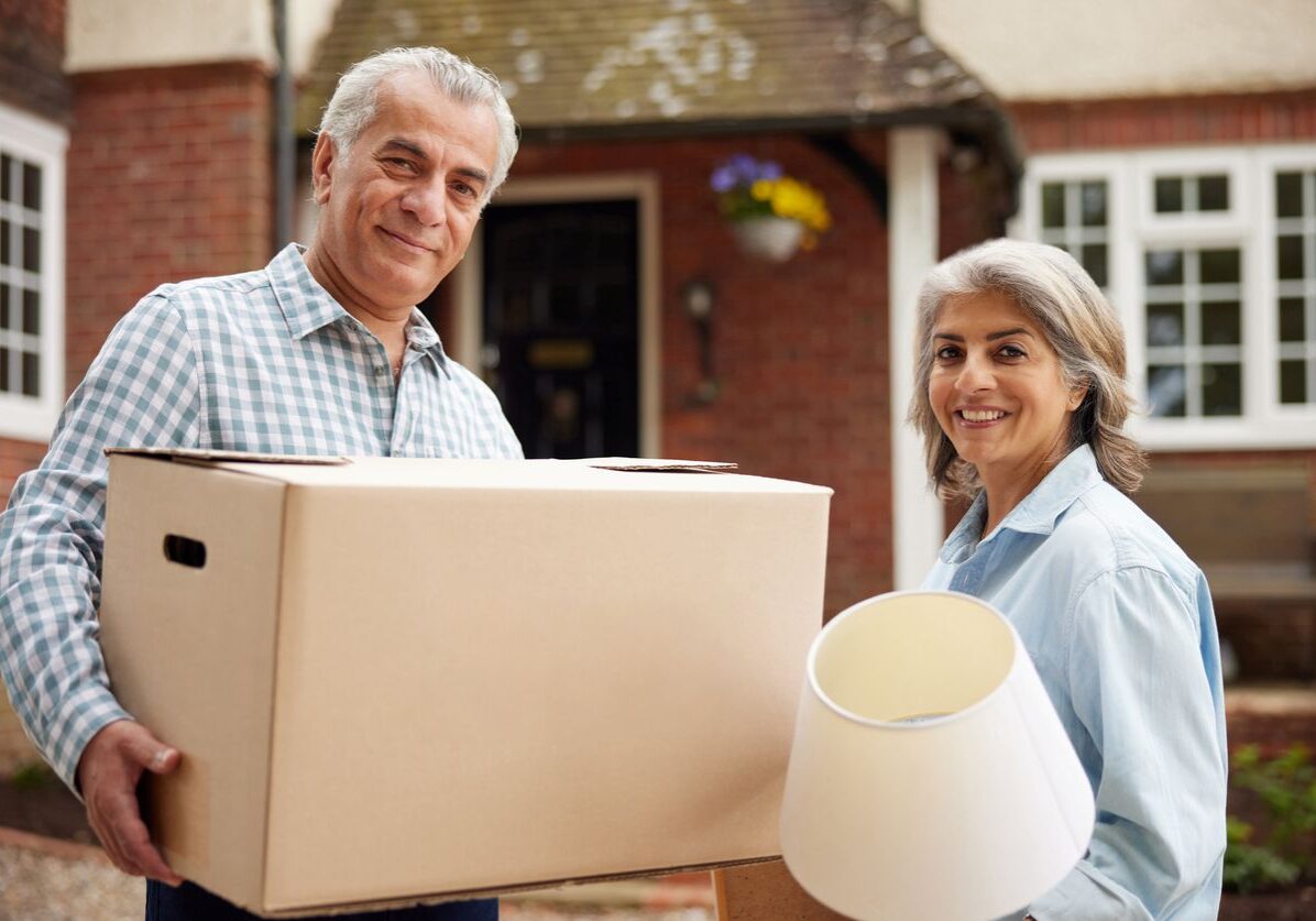 An older couple holding a box and lamp outside of a house.