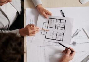 Two professionals reviewing an apartment floor plan and discussing furniture layout and room spacing.