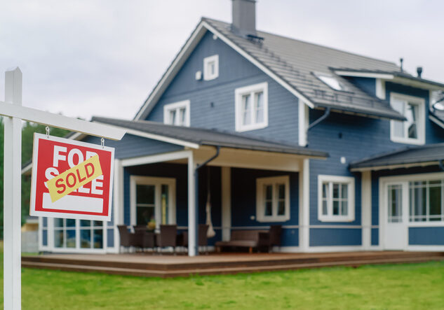 Red Property Sold Sign in a Garden in Front of a Big Blue Stylish House with Traditional Architecture. Hot Housing Market Concept with Residential Home in the Suburbs.