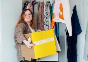 A woman smiles as she holds a box labeled “donations” in her closet.