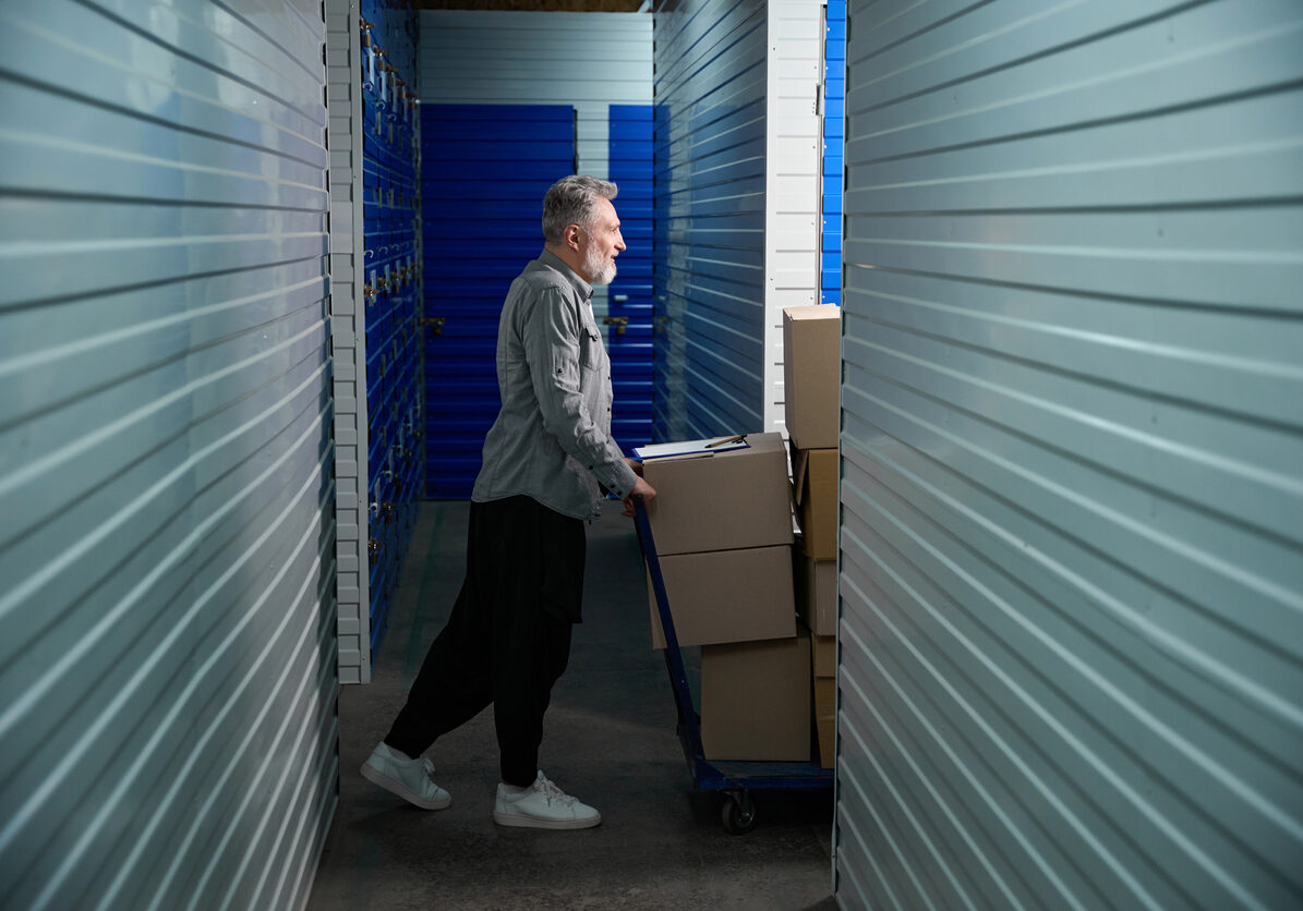 Man carrying large trolley filled with stacked boxes Adult man standing near big trolley with stacked boxes in storehouse and walking