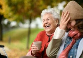 A granddaughter with her grandma sitting on a bench in autumn, laughing and drinking coffee.
